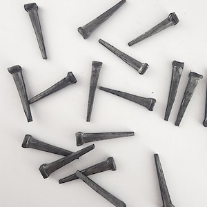 May include: A collection of dark gray cut nails scattered on a white surface. Each nail has a rectangular head and a tapered, pointed body. The nails vary in orientation, creating a random pattern.