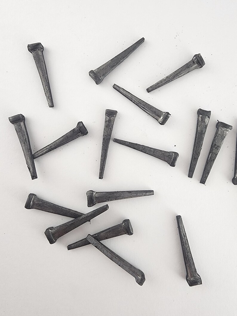 May include: A collection of dark gray cut nails scattered on a white surface. Each nail has a rectangular head and a tapered, pointed body. The nails vary in orientation, creating a random pattern.