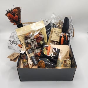 May include: A black cardboard box filled with various items including a small broom with orange flowers, a clear pouch of dried botanicals, a glass vial with a skull topper, a Samhain Mullein Torch, black candles, and a small Ouija board game.