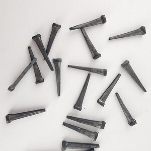 May include: A collection of dark gray cut nails scattered on a white surface. The nails have a rectangular head and a tapered, wedge-shaped body, ideal for woodworking and construction projects.
