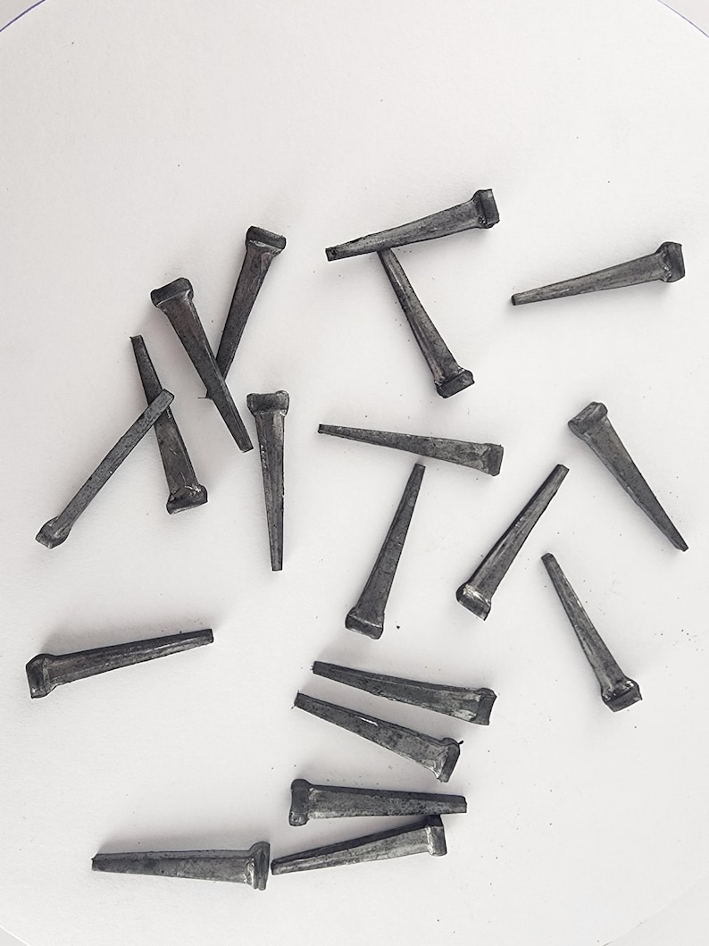 May include: A collection of dark gray cut nails scattered on a white surface. The nails have a rectangular head and a tapered, wedge-shaped body, ideal for woodworking and construction projects.
