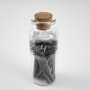 May include: A clear glass jar with a cork stopper, labeled "Coffin Nails" in a decorative font. The jar contains several gray, wedge-shaped objects. The label is black and white, and the jar is set against a white background.