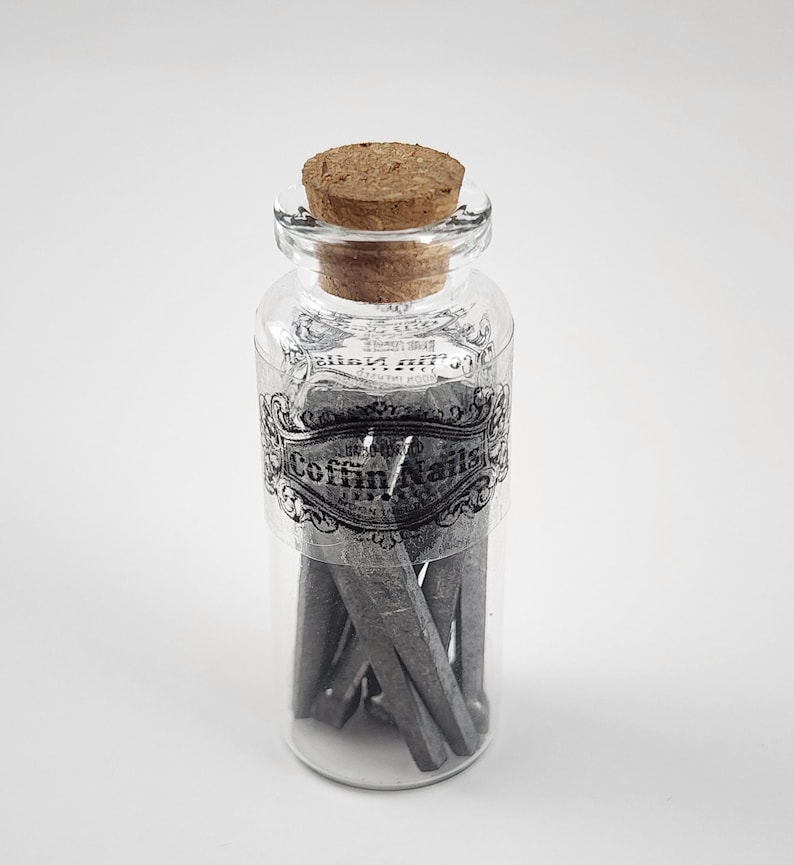 May include: A clear glass jar with a cork stopper, labeled "Coffin Nails" in a decorative font. The jar contains several gray, wedge-shaped objects. The label is black and white, and the jar is set against a white background.
