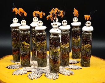 Puede incluir: Un conjunto de nueve viales de vidrio transparente rellenos con capas de hierbas, flores y especias secas. Cada vial está sellado con un tapón de cera blanca rematado con una pequeña figura de calavera y flores secas de color naranja.