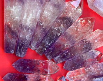May include: A collection of amethyst double terminated points, also known as double pointed crystals, with a clear to purple color gradient. The crystals are arranged in a fan shape on a red background.