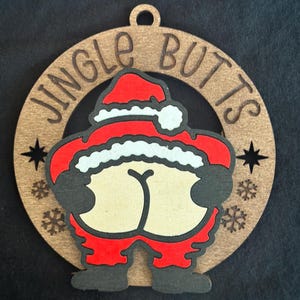 Op de afbeelding: Houten kerstversiering met een ontwerp van de kerstman en de tekst "JINGLE BUTTS". De cirkelvormige versiering heeft een uitgesneden ontwerp, met sneeuwvlok- en steraccenten. Gemaakt van hout.