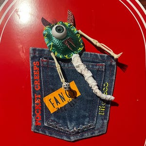 May include: A whimsical, handmade art piece featuring a denim pocket with the words "POCKET CREEPS" and "Bite Coo" in red and yellow. A green monster with a large eye and skeleton limbs is attached to the pocket, with a "FANG" tag.