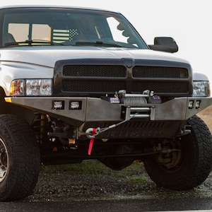 May include: A silver pickup truck with a black grille and a custom front bumper. The truck has large off-road tires and a winch. The background is a field with a sunset.