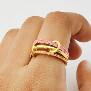 Könnte beinhalten: Ein goldfarbener Ring mit drei Bändern, von denen zwei mit kleinen rosa Edelsteinen besetzt sind. Die Bänder sind miteinander verschlungen und bilden ein einzigartiges Knotendesign. Der Ring wird an einem Finger vor weißem Hintergrund getragen.