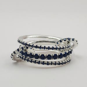 Op de afbeelding: Een zilveren ring met meerdere banden, elk bezet met kleine, donkerblauwe edelstenen. De banden zijn met elkaar verweven, waardoor een gelaagd en gestructureerd uiterlijk ontstaat. De ring staat tegen een effen witte achtergrond.