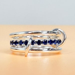 May include: A silver ring featuring three bands, each set with a line of small, dark blue gemstones. The ring has a distinctive design with a circular clasp and a small, vertical bar. The ring is displayed against a peach-coloured surface.