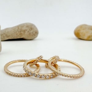 Op de afbeelding: Drie goudkleurige ringen met kleine, heldere edelstenen rond de banden. De ringen zijn in een licht overlappend patroon gerangschikt tegen een witte achtergrond. Twee gladde, lichtgekleurde stenen op de achtergrond.