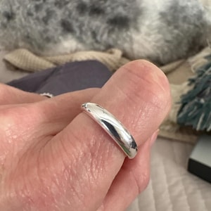 Könnte beinhalten: Ein polierter Silberring mit abgerundetem Band wird an einem Finger getragen. Der Ring reflektiert das Licht und erzeugt ein helles, glänzendes Aussehen. Der Ring hat ein schlichtes, klassisches Design.
