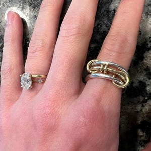 Peut inclure: Deux bagues sont présentées sur un doigt. L'une est une bague en or avec un grand diamant de taille ovale et deux petits diamants. L'autre bague est une bague en argent avec des accents dorés, avec un design entrelacé unique.