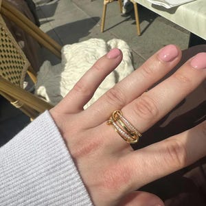 May include: A gold ring with an unusual design, featuring several interlocking bands. One band is set with small, sparkling stones. The ring is worn on a hand with pink nail polish, against a blurred outdoor backdrop.