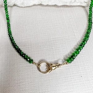 Puede incluir: Un collar de cuentas con pequeñas piedras preciosas verdes facetadas. El collar se asegura con un cierre de color dorado. Las cuentas son de un color verde intenso y el cierre tiene un diseño texturizado.