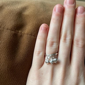 Könnte beinhalten: Ein silberner Ring mit drei baumelnden Herzanhängern. Der Ring wird an einem Finger getragen und zeigt das filigrane Design und die polierte Oberfläche. Die Herzen sind klein und gleichmäßig angeordnet und verleihen dem Schmuck Charme.
