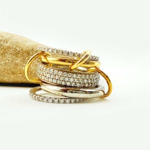 Può includere: Una collezione di anelli in oro e argento, alcuni incastonati con piccole pietre scintillanti. Gli anelli sono impilati, con alcuni anelli d'oro che si intrecciano attorno alla pila. Una pietra strutturata, color beige, sullo sfondo.