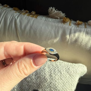 May include: A gold ring with a black enamel band and a central oval design. The oval features a blue gemstone surrounded by small, clear gemstones. The ring is held up against a soft, textured background.