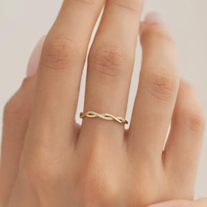 Peut inclure: Une bague dorée avec un motif tressé, ornée de petites pierres scintillantes. La bague est présentée sur un doigt, soulignant son aspect délicat et élégant. Le motif de la bague forme une boucle continue, suggérant l'infini.