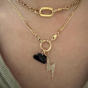 Op de afbeelding: Goudkleurige kettingkettingen. De onderste ketting heeft een zwarte wolkbedel en een met diamanten bezette bliksembedel. De bovenste ketting heeft een grote sluiting. De kettingen zijn gelaagd en worden om de nek gedragen.