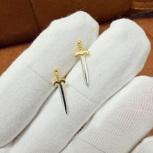 Op de afbeelding: Twee miniatuur zwaardhangers. De zwaarden hebben goudkleurige gevesten en zilverkleurige bladen. De hangers worden gepresenteerd op een witte stoffen achtergrond. De zwaarden zijn klein, waarschijnlijk voor sieraden.