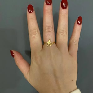 May include: A gold-colored ring in the shape of the letter "K" is worn on a finger. The hand has red nail polish. The ring is the focal point, set against a neutral background.