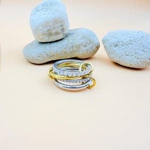 Op de afbeelding: Een stapelbare ringenset met zilveren en gouden banden. Eén ring is versierd met kleine, heldere edelstenen. De ringen zijn gerangschikt in een gelaagd ontwerp, wat een moderne en elegante uitstraling creëert. De achtergrond bevat lichte stenen.