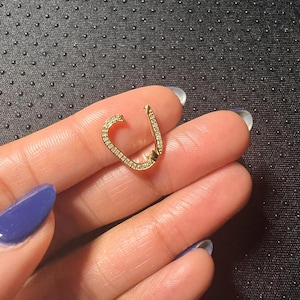 Op de afbeelding: Een goudkleurige oorbel in een uniek veiligheidsspeldontwerp, bezet met kleine, sprankelende edelstenen. De oorbel wordt in een hand gehouden, waarbij de ingewikkelde details en het licht dat weerkaatst op de edelstenen worden benadrukt.