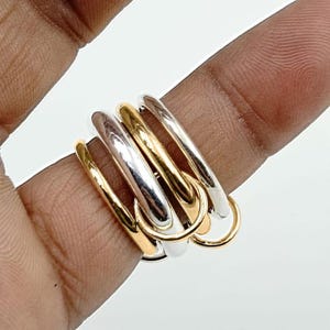Könnte beinhalten: Ein mehrfarbiger Ring mit verbundenen Bändern in Gold, Silber und Roségold. Der Ring wird auf einem Finger gezeigt, wodurch sein geschichtetes Design und sein metallischer Glanz hervorgehoben werden. Die Bänder sind glatt und poliert.