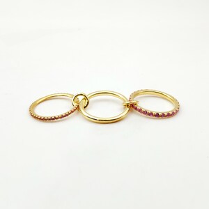 Peut inclure: Un ensemble de bagues dor&eacute;es comprenant trois anneaux reli&eacute;s. Deux des anneaux sont orn&eacute;s de petites pierres roses. Les anneaux sont reli&eacute;s par un petit connecteur circulaire, cr&eacute;ant un design de bijoux unique.