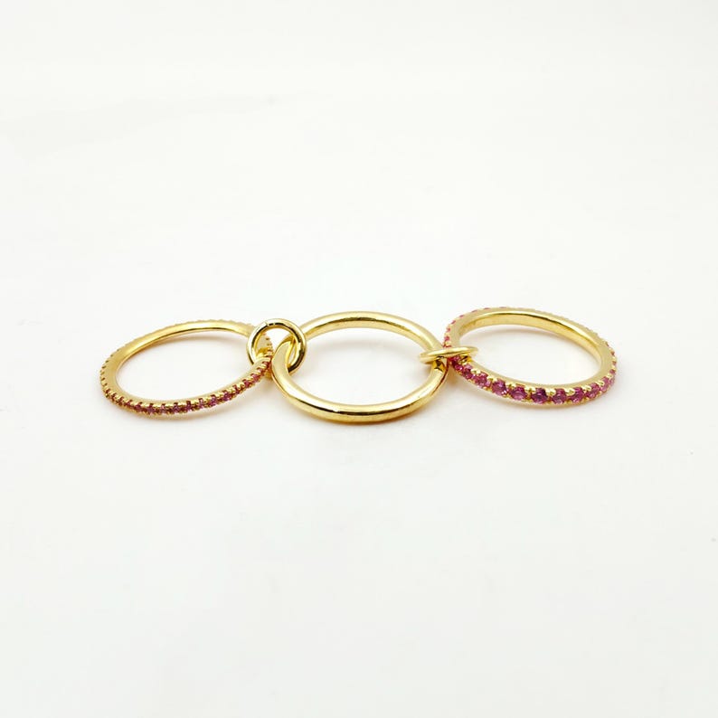 Peut inclure: Un ensemble de bagues dor&eacute;es comprenant trois anneaux reli&eacute;s. Deux des anneaux sont orn&eacute;s de petites pierres roses. Les anneaux sont reli&eacute;s par un petit connecteur circulaire, cr&eacute;ant un design de bijoux unique.