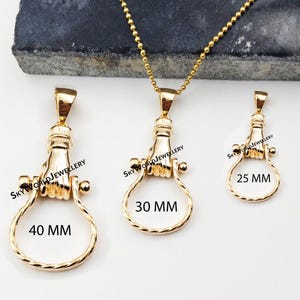 Puede incluir: Tres colgantes de joyería dorados con forma de mano que sostiene un anillo circular trenzado. Los colgantes están etiquetados como 40 MM, 30 MM y 25 MM. Un colgante está unido a una cadena de bolas dorada. El texto SKYWORLDJEWELLERY es visible en cada uno.