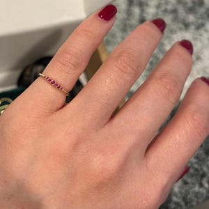 May include: A gold ring with a row of small, red gemstones. The ring is delicate and sits on a finger. The gemstones are evenly spaced and add a pop of color to the gold band. Jewelry.