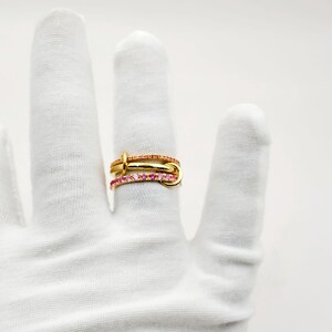 Peut inclure: Trois bagues dor&eacute;es sont pr&eacute;sent&eacute;es sur un doigt gant&eacute; de blanc. La bague sup&eacute;rieure est orn&eacute;e de pierres pr&eacute;cieuses orange, tandis que la bague inf&eacute;rieure pr&eacute;sente des pierres pr&eacute;cieuses roses. Les bagues sont imbriqu&eacute;es, cr&eacute;ant un design unique et &eacute;l&eacute;gant.