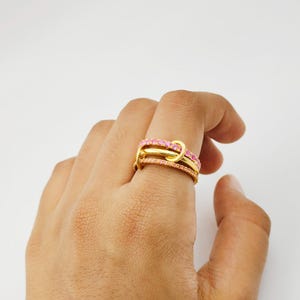 Op de afbeelding: Een goudkleurige ring met drie banden, waarvan twee bezet zijn met kleine roze edelstenen. De banden zijn met elkaar verweven en creëren een uniek knoopontwerp. De ring wordt op een vinger gedragen tegen een witte achtergrond.