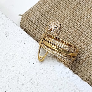 Puede incluir: Un anillo dorado con múltiples bandas, algunas incrustadas con pequeñas piedras transparentes. Un gran imperdible dorado sujeta las bandas, con una parte superior redonda adornada con gemas. El anillo está sobre una superficie texturizada.