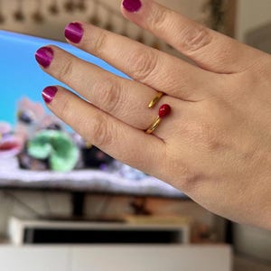 Puede incluir: Un anillo dorado con un diseño de cabeza de cerilla roja. El anillo abierto tiene la cabeza de la cerilla en el dedo. La mano que lleva el anillo tiene las uñas pintadas de color magenta.