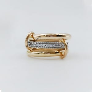 Op de afbeelding: Een goudkleurige ring met een centrale band van kleine, heldere stenen. De ring heeft een meerlagig ontwerp met gebogen, gladde banden en verbindingslussen. De tekst "Buzzlebeejewels" is zichtbaar op de ring.