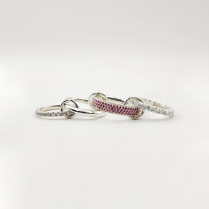 May include: Three silver rings are interlocked, each with a unique design. One ring is adorned with small, clear stones, another with a band of pink stones, and the third is a simple silver band. The rings are displayed on a white surface.