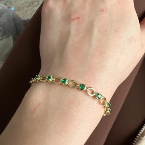 以下が含まれることがあります： エメラルドグリーンの宝石が付いたゴールドのブレスレット。ブレスレットは、楕円形のリンクと、均等に配置された丸く鮮やかな緑色の石を備えた繊細なチェーンデザインが特徴です。ブレスレットは手首に着用されています。