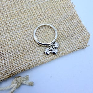 Könnte beinhalten: Ein silberner Ring mit drei baumelnden Herzanhängern. Der Ring liegt auf einem beigen Jutebeutel mit Kordelzug. Das Design des Rings ist schlicht und elegant, geeignet für den täglichen Gebrauch.