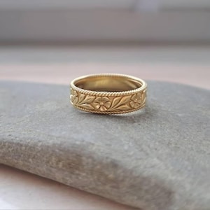 May include: A gold-colored ring with a floral pattern. The ring features a repeating design of flowers and leaves, bordered by a rope-like texture. The ring is set on a gray stone surface.