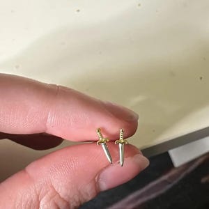 Op de afbeelding: Een paar kleine, zilverkleurige dolk oorbellen met goudkleurige handvatten. De oorbellen worden getoond tegen een neutrale achtergrond.