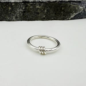 Puede incluir: Un anillo de plata con un diseño único. El anillo presenta una banda lisa y redondeada con tres pequeñas bandas texturizadas agrupadas en la parte delantera. El anillo está sobre una superficie blanca.