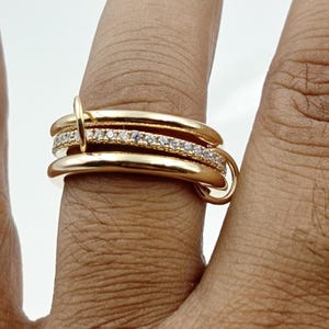May include: A gold-colored ring with three stacked bands. The middle band is encrusted with small, clear gemstones. The ring is connected by a small, circular clasp. The ring is on a finger.