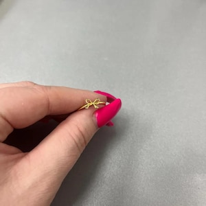 May include: A gold-coloured ring with a bow design. The ring is held between fingers with bright pink nail polish, set against a grey background. The bow is the focal point of the ring.