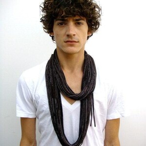 May include: A person wearing a white V-neck t-shirt and a dark gray scarf. The scarf is long and looped around the neck. The person has dark curly hair.