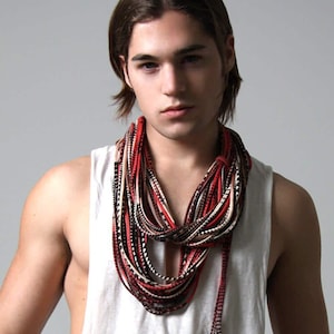 Può includere: Una collana a più fili nei colori rosso, nero e crema. La collana è realizzata con sottili fili simili a tessuto. La collana è indossata sopra una canotta bianca.