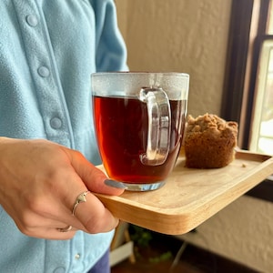 Può includere: Un vassoio di legno con una tazza di vetro trasparente di tè scuro e un muffin su una superficie di legno.